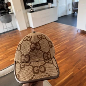 Beige keps med GG-mönster - Snygg beige keps från Gucci med det ikoniska GG-mönstret i brunt. Kepsen har en justerbar rem baktill för perfekt passform. Perfekt för att ge din outfit en lyxig touch.