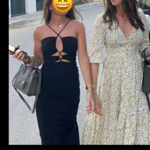 Svart långklänning med slits - Elegant svart långklänning med smala axelband och en slits baktill. Klänningen har en unik detalj framtill med utskärningar och knytning, vilket ger en modern och stilren look.