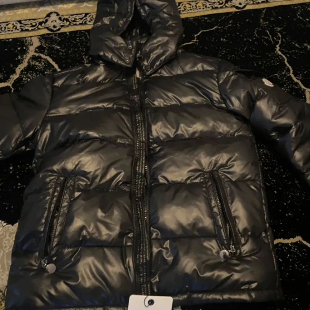Säljer en snygg svart dunjacka från Moncler med huva och dragkedja. Jackan har en glansig finish och praktiska fickor med knappar. Perfekt för kyliga dagar när du vill hålla stilen. 🖤passar L och M. Takit.