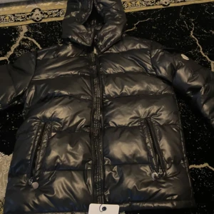  Moncler maya - Säljer en snygg svart dunjacka från Moncler med huva och dragkedja. Jackan har en glansig finish och praktiska fickor med knappar. Perfekt för kyliga dagar när du vill hålla stilen. 🖤passar L och M