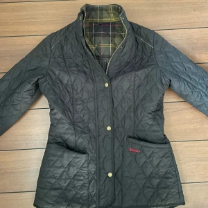 Svart quiltad jacka från Barbour - Säljer en stilren svart quiltad jacka från Barbour med klassiskt tartanmönstrat foder. Jackan har knappar framtill och två praktiska fickor. Perfekt för kyligare dagar!