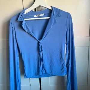 Blå långärmad topp från NA-KD - Snygg blå långärmad topp från NA-KD med knytning framtill. Perfekt för en stilren look. Toppen har en krage och är tillverkad i ett mjukt material.