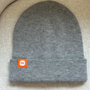 Grå mössa från Xiaomi - Snygg grå mössa från Xiaomi i mjukt akrylmaterial. Den har en enkel design med en uppvikt kant och en liten orange logga framtill. 