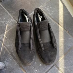 Svarta slip-on skor från Arigato - Snygga svarta slip-on skor från Arigato i mocka. Skorna har en stilren design med elastiska paneler för enkel på- och avtagning. Perfekta för en avslappnad men stilfull look.