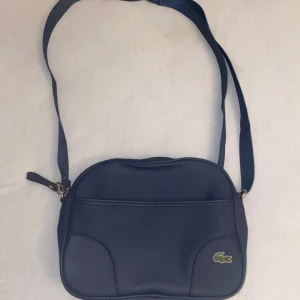 Blå axelväska från Lacoste - Snygg blå axelväska från Lacoste med justerbar rem och dragkedja. Perfekt för att bära dina dagliga nödvändigheter med stil. Den ikoniska krokodilloggan pryder framsidan.