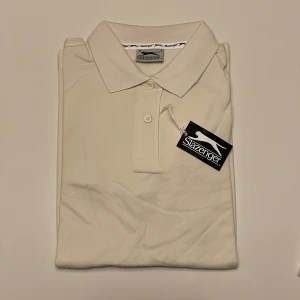 Vit pikétröja från Slazenger - Säljer en helt ny stilren vit pikétröja från Slazenger. Tröjan har en klassisk krage med två knappar och korta ärmar. Perfekt för en sportig look eller en avslappnad dag. 🏌️‍♂️