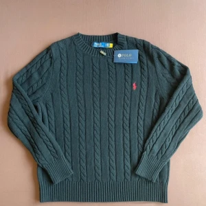 Grå stickad tröja från Polo Ralph Lauren - Säljer en snygg grå stickad tröja från Polo Ralph Lauren med kabelstickat mönster. Tröjan har en broderad röd logotyp på bröstet och långa ärmar. Perfekt för kyligare dagar!