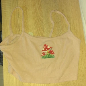 Beige ribbad linnetopp från Shein - Säljer en söt beige ribbad linnetopp från Shein med broderade svampar på framsidan. Toppen har smala axelband och är perfekt för en avslappnad stil. Passar bra till jeans eller shorts för en cool look.