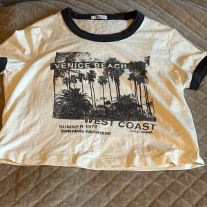 Vit t-shirt med tryck från Ardene i New York - Vit-shirt från Ardene med ett svart tryck av palmer och texten 'Venice Beach West Coast'. T-shirten har svarta kanter vid hals och ärmar. 