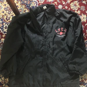 Svart vindjacka från Adidas - Säljer en svart vindjacka från Adidas med FCN-logga på bröstet. Jackan har dragkedja och elastiska ärmslut. Perfekt för blåsiga dagar har små fläckar på bröstet syns dock inte så tydligt .