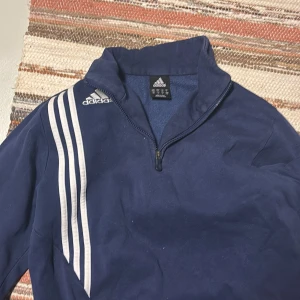 Blå tröja med dragkedja från Adidas - Säljer en blå tröja från Adidas med dragkedja och de klassiska tre vita ränderna på ena sidan. Perfekt för en sportig look eller avslappnad stil. Tröjan har långa ärmar och en hög krage.