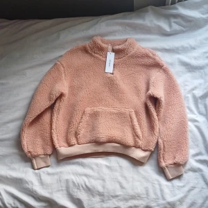 Rosa teddy tröja - Mysig rosa teddyfleece tröja från Size & Needle med lång ärm och en praktisk magficka. Perfekt för kyliga dagar med sin mjuka och varma design. Tröjan har ribbade muddar vid ärmslut och nederkant för extra komfort.