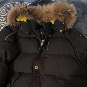 Mooseknuckles  - Säljer en svart guld Mooseknuckles med snygga pälsdetaljer på huvan. Jackan har dragkedja och knappar framtill samt praktiska fickor. Perfekt för kalla dagar med sin quiltade design och långa ärmar. 