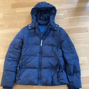 Mörkblå pufferjacka från GANT - Säljer en snygg mörkblå jacka från GANT med huva och dragkedja. Den är 9/10 i skick inga skador eller fläckar riktigt bra skick!