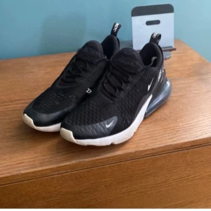 Svarta Nike Air Max 270 - Säljer ett par svarta Nike Air Max 270 med vit sula och synlig luftkudde i hälen. Skorna har en stilren design med snörning och små vita detaljer. Perfekta för en sportig look. Se bilder för egen uppfattning om skick.