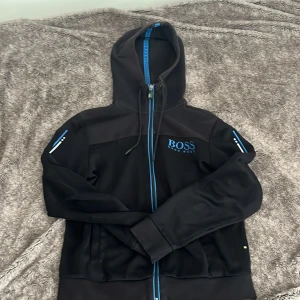 Svart hoodie från Hugo Boss - Snygg svart hoodie från Hugo Boss med blå detaljer och dragkedja. Den har en broderad logga på bröstet och en huva med snörning. Perfekt för en stilren och bekväm look.