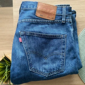 Blå Levis 501 jeans - Klassiska blå Levis 501 jeans med rak passform och knappgylf. Perfekta för en avslappnad stil. Midjan är normalhög och byxorna har den ikoniska Levis-lappen på baksidan.