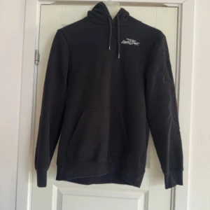 Svart hoodie med tryck - Säljer en svart hoodie med ett unikt tryck på ärmen och ryggen. Hoodien har en klassisk design med dragsko i huvan och en stor magficka framtill. Perfekt för en avslappnad stil.