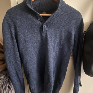 Blå stickad tröja  - Säljer en snygg blå stickad tröja. Tröjan har en hög krage med knappar och långa ärmar. Perfekt för kyligare dagar. Passar bra till både jeans och chinos.