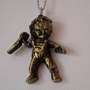 Halsband med figurhänge Chucky  - Unikt halsband med ett hänge i form av en figur i antikguld. Figuren Chucky detaljer som en kniv i handen och en overall. Perfekt för den som gillar lite annorlunda smycken.