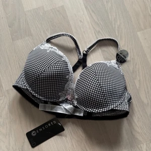 Svartvit push-up bh med spetsdetaljer - Säljer en snygg svartvit push-up bh från Censored med avtagbara inlägg. Den har ett rutigt mönster och fina spetsdetaljer vid kuporna. En söt rosett pryder framsidan. Perfekt för att ge extra lyft och form. Aldrig använd! Storlek 85D