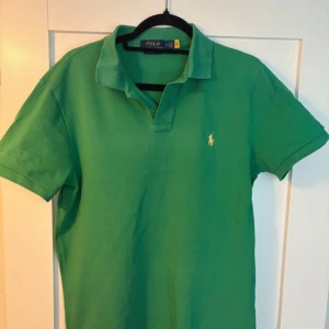 Grön kortärmad pikétröja från Ralph Lauren - Säljer en snygg grön pikétröja från Ralph Lauren. Tröjan är kortärmad och har en klassisk krage med knappslå. Den är tillverkad i bomull och har en liten broderad logga på bröstet. Perfekt för en stilren look!