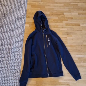 Mörkblå hoodie från sail racing - Säljer en snygg mörkblå hoodie från Peak Performance med dragkedja framtill. Perfekt för kyliga dagar med sin bekväma passform och långa ärmar. Huvan ger extra värme och stil. Passar bra till både jeans och träningsbyxor.