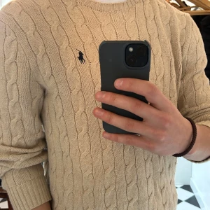 Beige stickad tröja från Ralph Lauren - Säljer en stilren beige stickad tröja från Ralph Lauren med kabelstickat mönster. Tröjan har långa ärmar och en liten broderad logotyp på bröstet. Perfekt för en avslappnad och elegant look. Fortfarande mjuk och sitter perfekt på mig som är 180 cm!