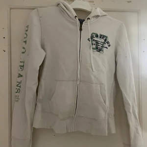 Vit hoodie från Polo Jeans Co. Ralph Lauren - Säljer en vit hoodie från Polo Jeans Co. Ralph Lauren med dragkedja och tryck på bröstet och ärmen. Tillverkad i en bomulls- och polyesterblandning. Perfekt för en avslappnad stil. 🧢