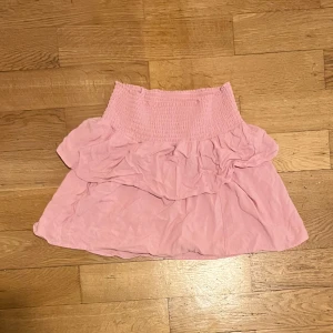 Rosa kortkjol med volang - Söt rosa kortkjol med volangdetaljer. Perfekt för varma dagar och ger en lekfull touch till din outfit. Den har en elastisk midja för bekväm passform.