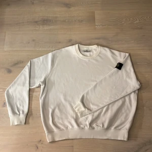 Vit sweatshirt från Stone Island - Säljer en stilren vit sweatshirt från Stone Island. Tröjan har en klassisk rund halsringning och en ikonisk logotyp på ärmen. Perfekt för en avslappnad och trendig look.