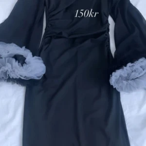 Svart klänning med volangärmar - Snygg svart klänning med långa ärmar och volangdetaljer vid ärmsluten. Perfekt för en elegant look. Klänningen har en omlott-design som ger en fin passform.