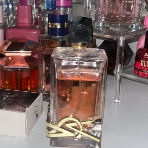 Parfym från Yves Saint Laurent - Elegant parfymflaska från Yves Saint Laurent med en rektangulär form och en stilren svart kork. Flaskan är dekorerad med ett guldigt YSL-emblem och innehåller en ljusrosa doftvätska. Perfekt för den som älskar lyxiga och sofistikerade dofter.