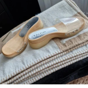 Clearheels - Snygga träskor från Kaltur med en modern twist. De har en transparent rem över foten och en klassisk träfärgad sula. Perfekta för en stilren look.
