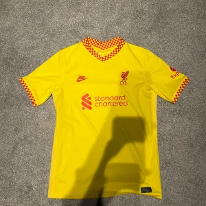 Liverpool 21/22 tredje ställ storlek 158-170 fotbollströja från Nike - Säljer en Liverpool fotbollströja från Nike med röda detaljer och rutigt mönster vid krage och ärmslut. Tröjan har korta ärmar och är perfekt för fotbollsfans. Den har tryck av sponsorer och klubbmärke på bröstet.