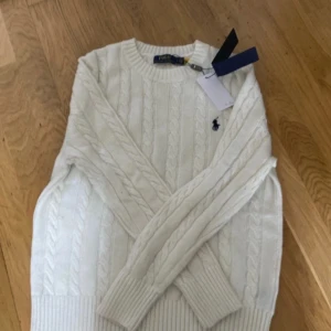 Vit stickad tröja från Ralph Lauren - Säljer en stilren vit stickad tröja från Ralph Lauren med kabelstickat mönster och den klassiska loggan på bröstet. Tröjan har långa ärmar och ribbstickade muddar vid ärmslut och nederkant. Perfekt för kyliga dagar!