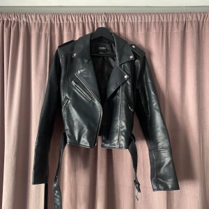 Svart skinnjacka - Snygg svart skinnjacka från Pull & Bear med dragkedja och bälte. Jackan har en croppad passform och långa ärmar. Perfekt för en cool look!