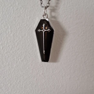 Svart kistformat halsband med kors - Unikt halsband med en svart kistformad berlock och ett silverfärgat kors i mitten. Kedjan är också silverfärgad och ger en stilren look. Perfekt för att ge en edgy touch till din stil.