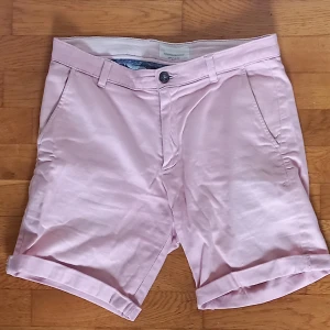 Rosa shorts från Tailored Originals - Snygga rosa shorts från Tailored Originals med knapp och dragkedja framtill. De har två sidofickor och två bakfickor med knappar. Perfekta för en avslappnad stil.