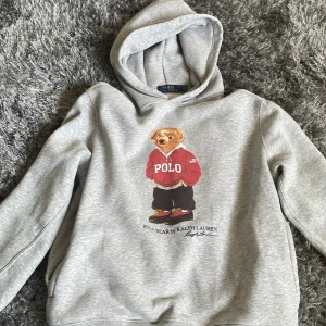 Grå hoodie från Ralph Lauren med Polo Bear - Säljer en grå hoodie från Ralph Lauren med det ikoniska Polo Bear-motivet på framsidan. Tröjan har en bekväm passform och är perfekt för en avslappnad stil. Den har långa ärmar och en klassisk huva. Perfekt för dig som älskar stil och komfort!