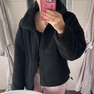 Svart teddyjacka från c.w - Mysig svart teddyjacka från C.W. med dragkedja framtill. Jackan har en hög krage och långa ärmar, perfekt för kyliga dagar. Den är tillverkad i ett mjukt och fluffigt material som håller dig varm och bekväm.