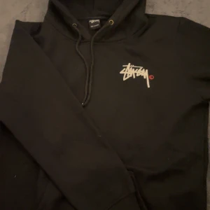Svart hoodie från Stüssy - Säljer en svart hoodie från Stüssy med deras ikoniska logga tryckt på bröstet och ryggen. Hoodien har en klassisk design med dragsko i huvan och ribbade muddar vid ärmslut och nederkant. Perfekt för en avslappnad stil. Passar bra för L, Mer baggy för M. Använd 1 gång så helt ny. 