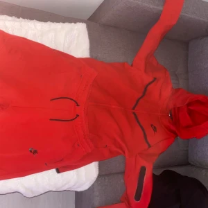 Röd Nike tech fleece - Säljer en röd hoodie från Nike med dragkedja och huva. Den har svarta detaljer och en ficka framtill. Perfekt för en sportig look eller avslappnade dagar.
