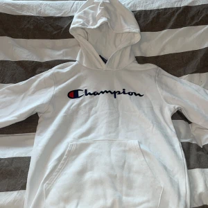 Vit hoodie från Champion - Säljer en vit hoodie från Champion med klassisk logga på bröstet. Den har en stor ficka framtill och en bekväm huva. Perfekt för en avslappnad stil.