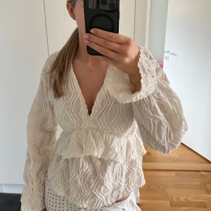 Vit blus med volangdetaljer - Säljer en söt vit blus med volangdetaljer och v-ringning. Blusen har långa ärmar och ett strukturerat tyg, säljer då jag beställde två storlekar och det är försent att skicka tillbaka😇