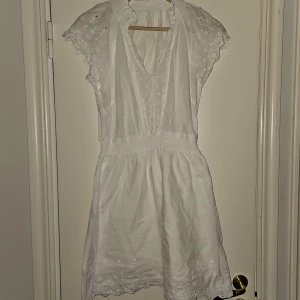 Vit klänning nytt - Vit klänning med broderade detaljer. Klänningen har en v-ringning och resår i midjan. Den har korta ärmar med broderade kanter och en broderad kant nedtill. Klänningen har underkjol