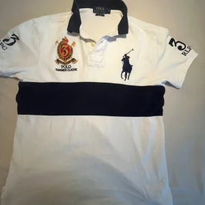 Vit och marinblå pikétröja från Ralph Lauren - Säljer en stilren vit pikétröja från Ralph Lauren med marinblå detaljer. Tröjan har en broderad logga på bröstet och en bred marinblå rand över magen. Perfekt för en klassisk och sportig look.