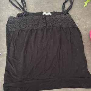 Svart linne från Vila - Säljer ett svart linne från Vila med spetsdetaljer upptill och knappar framtill. Linnet har justerbara axelband och en elegant design som passar perfekt för olika tillfällen.