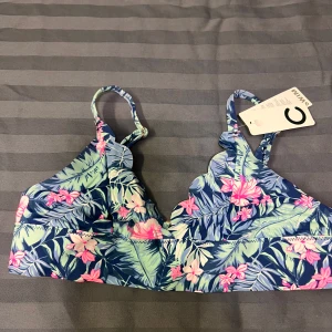 Blommig bikiniöverdel från Cubus - Snygg bikiniöverdel från Cubus med tropiskt blommönster i blått, grönt och rosa. Den har justerbara axelband och en praktisk spänne baktill. Perfekt för en dag på stranden eller vid poolen!