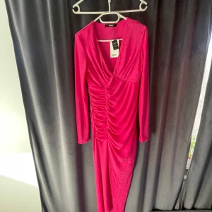 Rosa långklänning från Bik Bok - Snygg rosa långklänning från Bik Bok med v-ringning och rynkade detaljer framtill. Klänningen har långa ärmar och är perfekt för en elegant kväll. Med slits.Materialet ger en lyxig känsla och den smickrande passformen framhäver figuren.